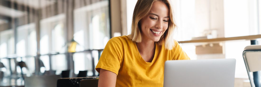 Photo of joyful nice woman using laptop and smiling while sitting at table in open-plan office