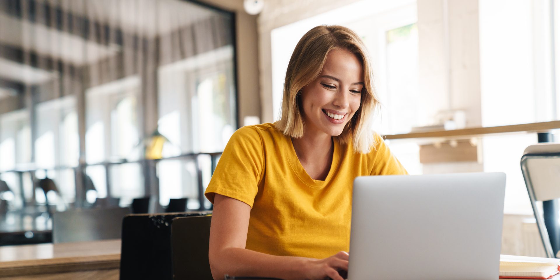 Photo of joyful nice woman using laptop and smiling while sitting at table in open-plan office