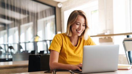 Photo of joyful nice woman using laptop and smiling while sitting at table in open-plan office